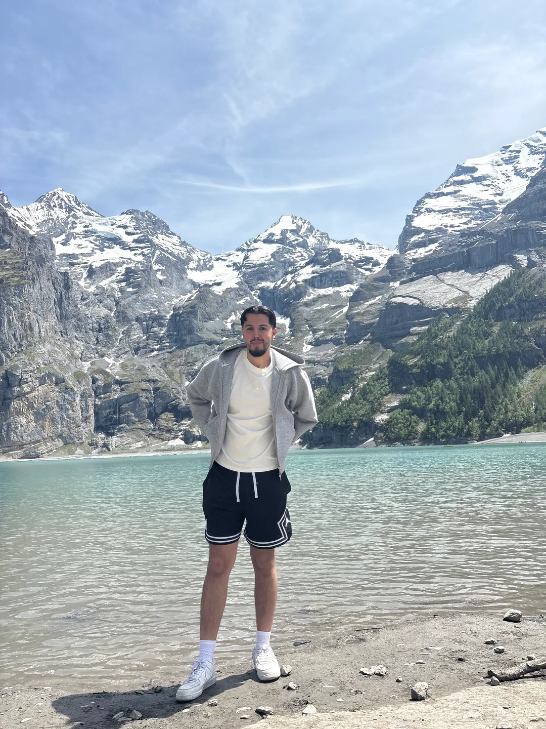 Ilyas standing by a turquoise mountain lake, snow-capped peaks rising behind him
