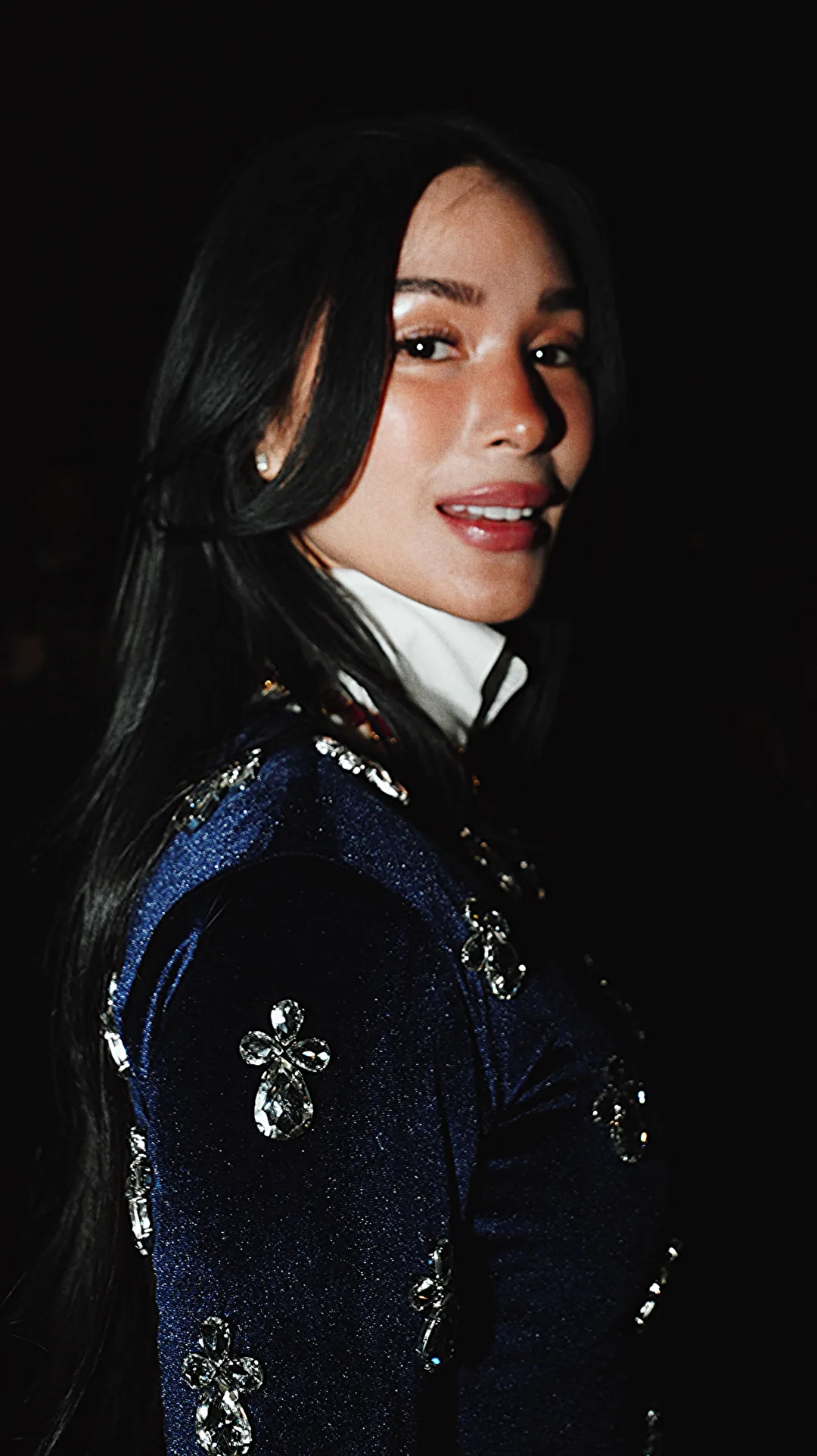 Portrait of a woman with long dark hair in a blue embellished jacket and white collar against darkness