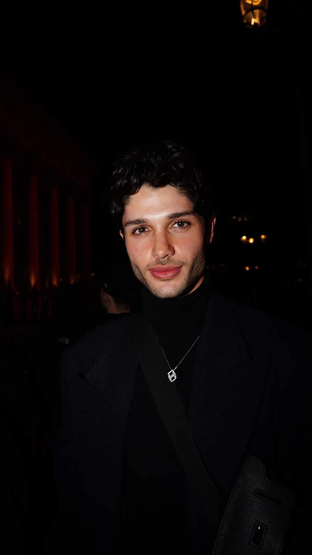 Low-light portrait of a man with dark curly hair in a black blazer and turtleneck with a fine necklace