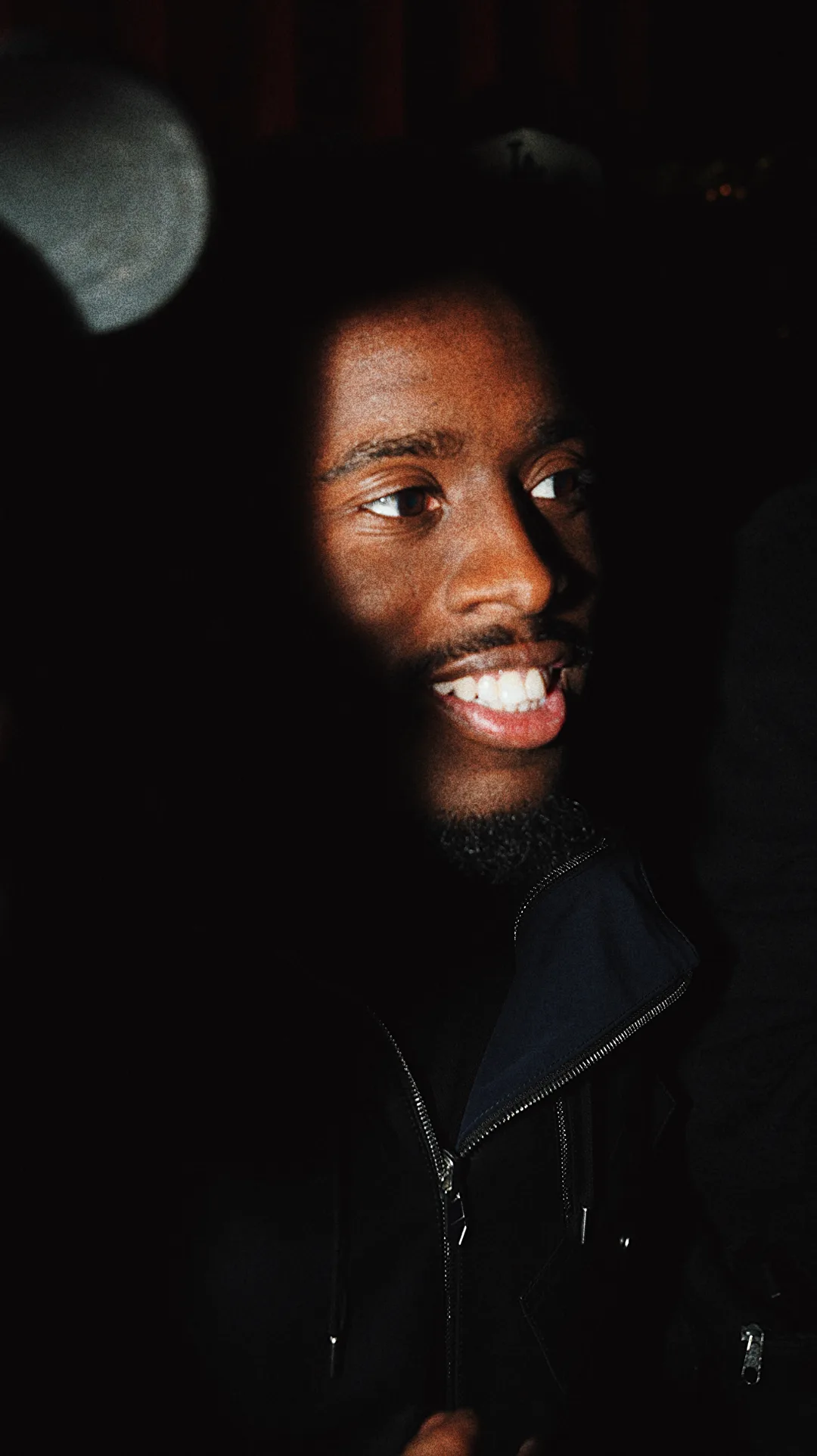 Low-light portrait of a smiling man with a beard in a dark jacket against a deep red background