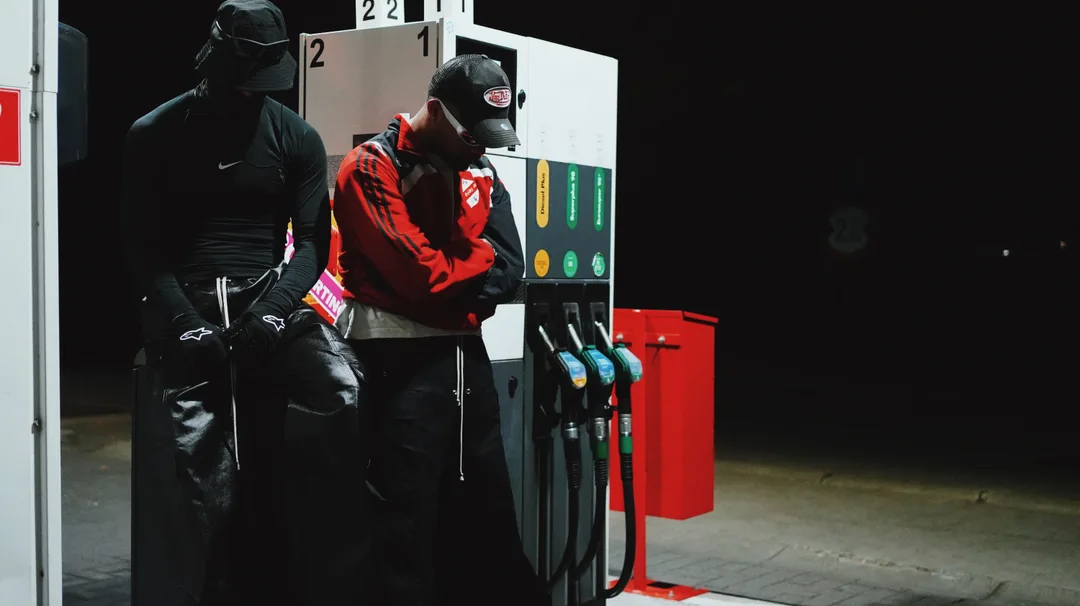 Two figures in streetwear at a petrol station at night, lit cinematically against the dark