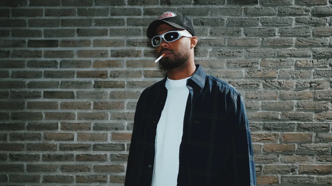 A man in a dark overshirt and cap standing against a brick wall