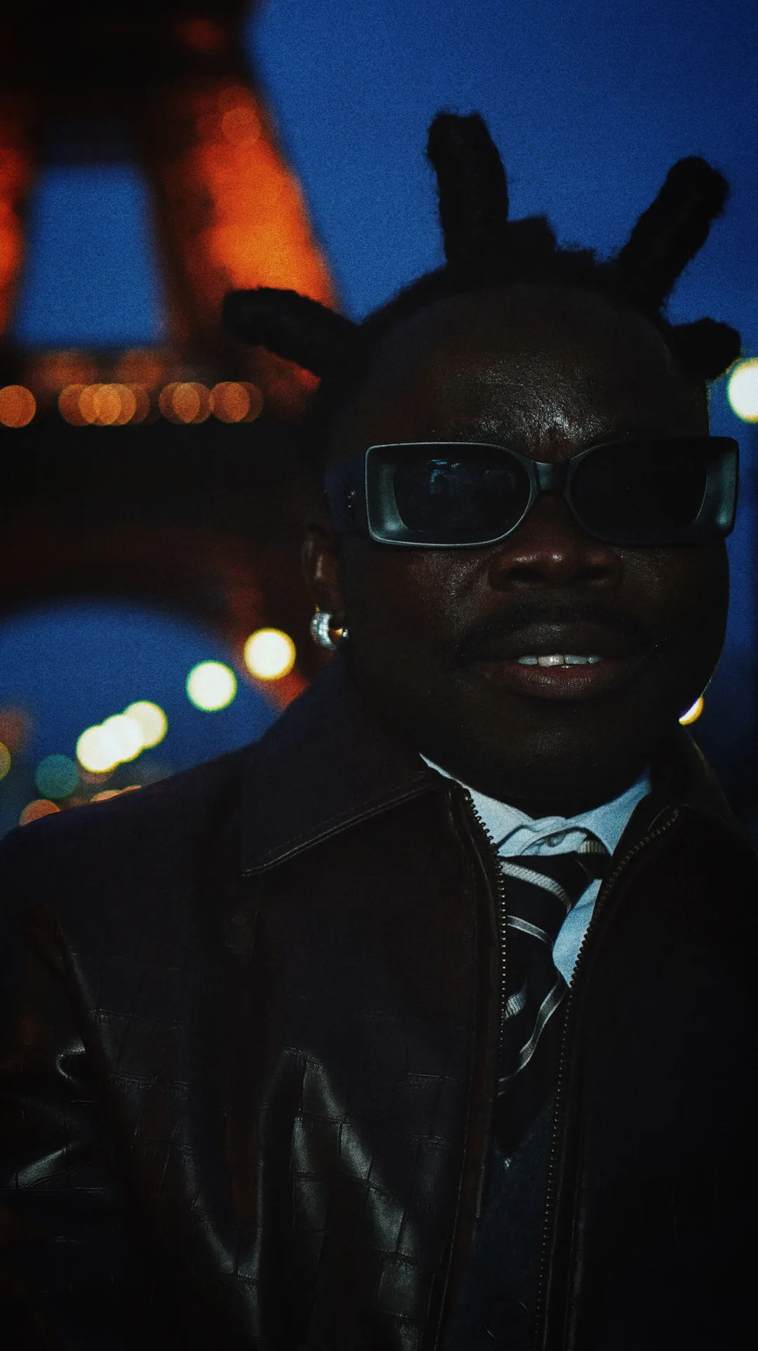 A man in a leather jacket and sunglasses in front of the illuminated Eiffel Tower at night