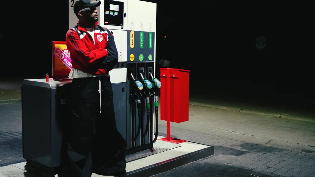 A figure in a red jacket leaning against a petrol pump at night