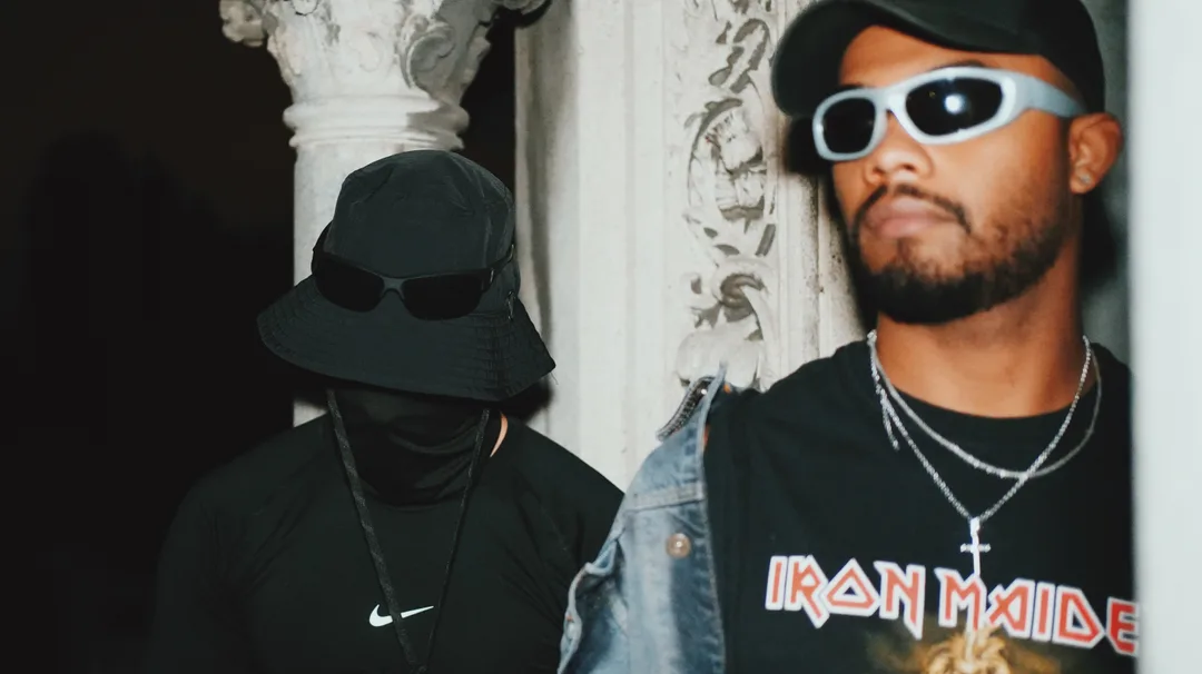Two figures, one masked, in band merchandise and denim against an ornate column