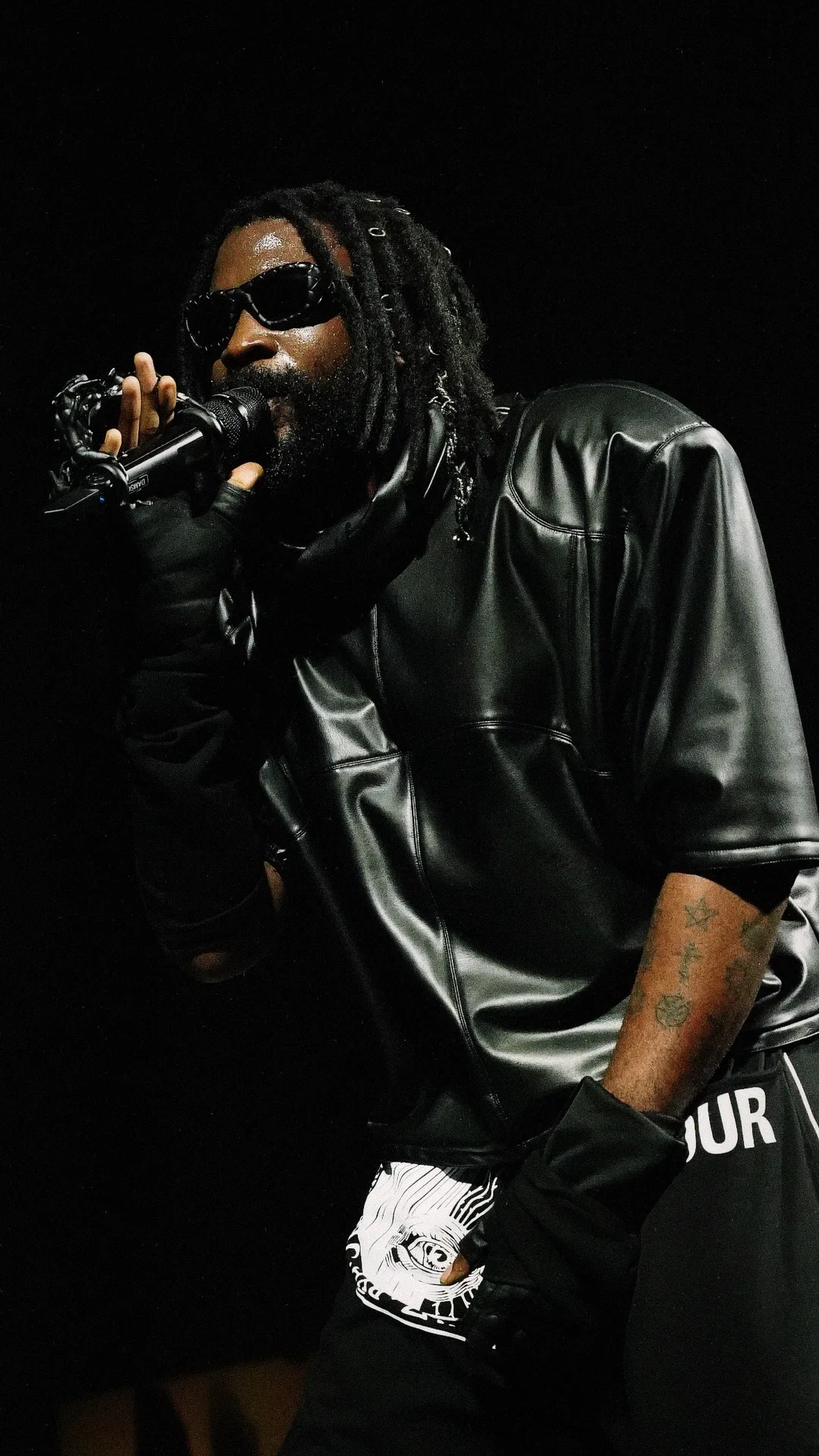 A rapper performing live with a microphone, wearing sunglasses and a leather tour jersey