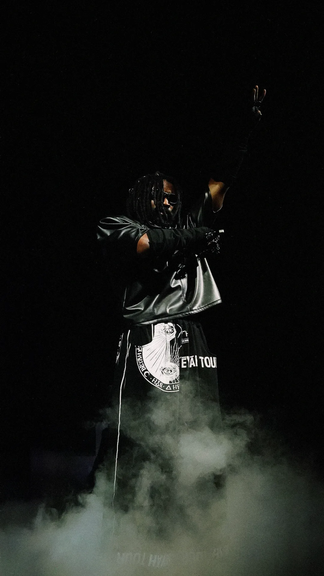 A performer on a smoke-filled stage with arms raised, lit from behind in darkness