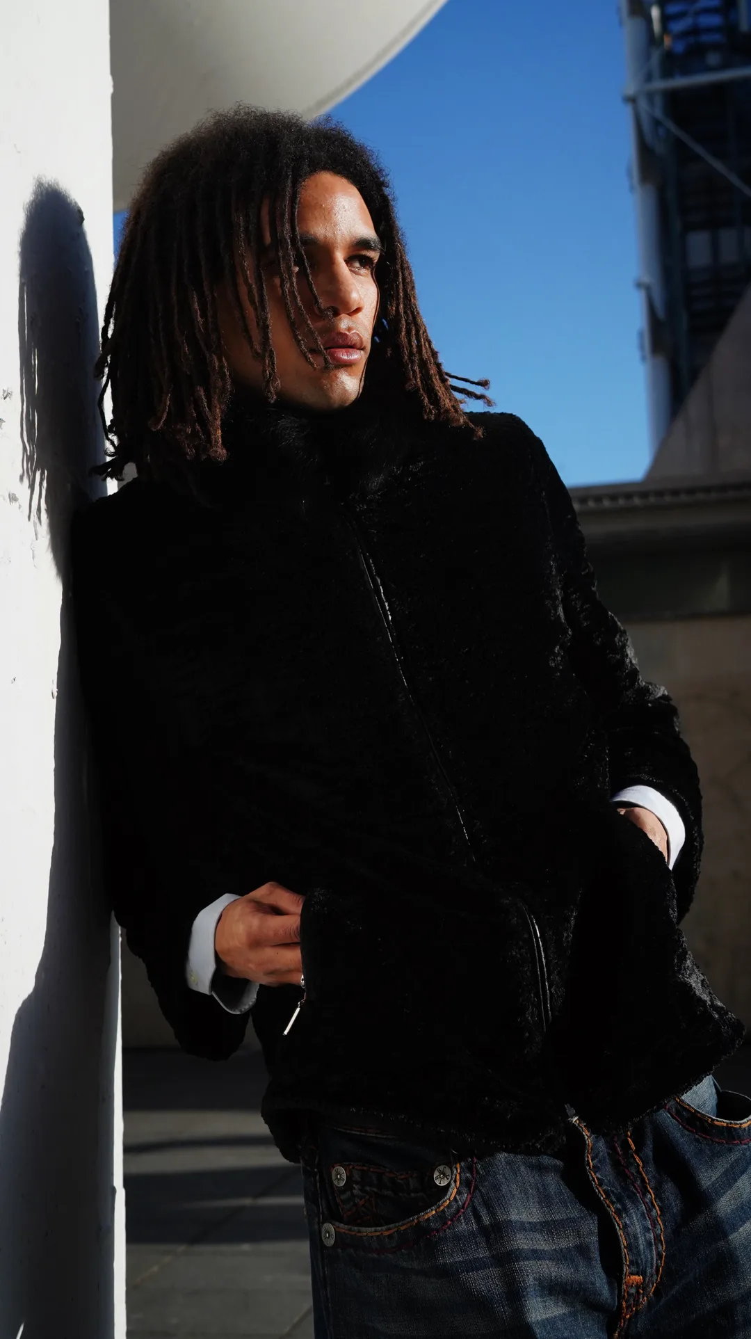 Portrait of a man with long dreadlocks in a black jacket against a white wall under blue sky