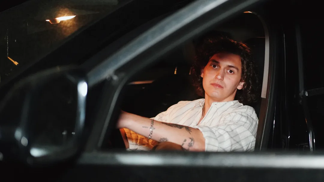 A man with curly hair at the wheel of a car at night